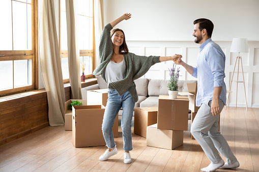 Happy young wife and husband dancing in modern living room with cardboard boxes with belongings, excited family celebrating moving day, satisfied customers relocating into new apartment, mortgage